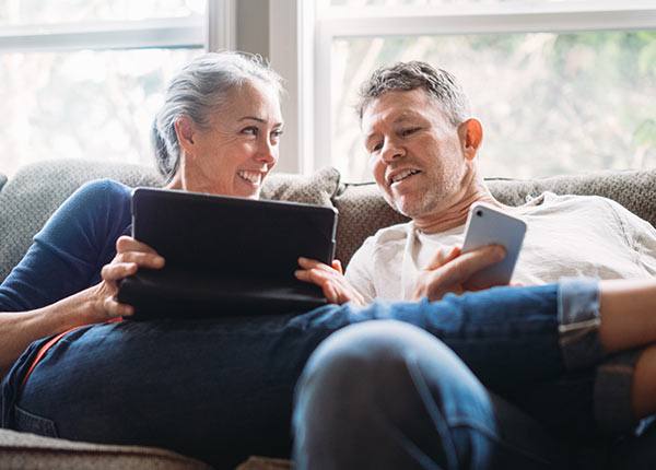 older couple sitting together on a couch each with their own devices and smiling